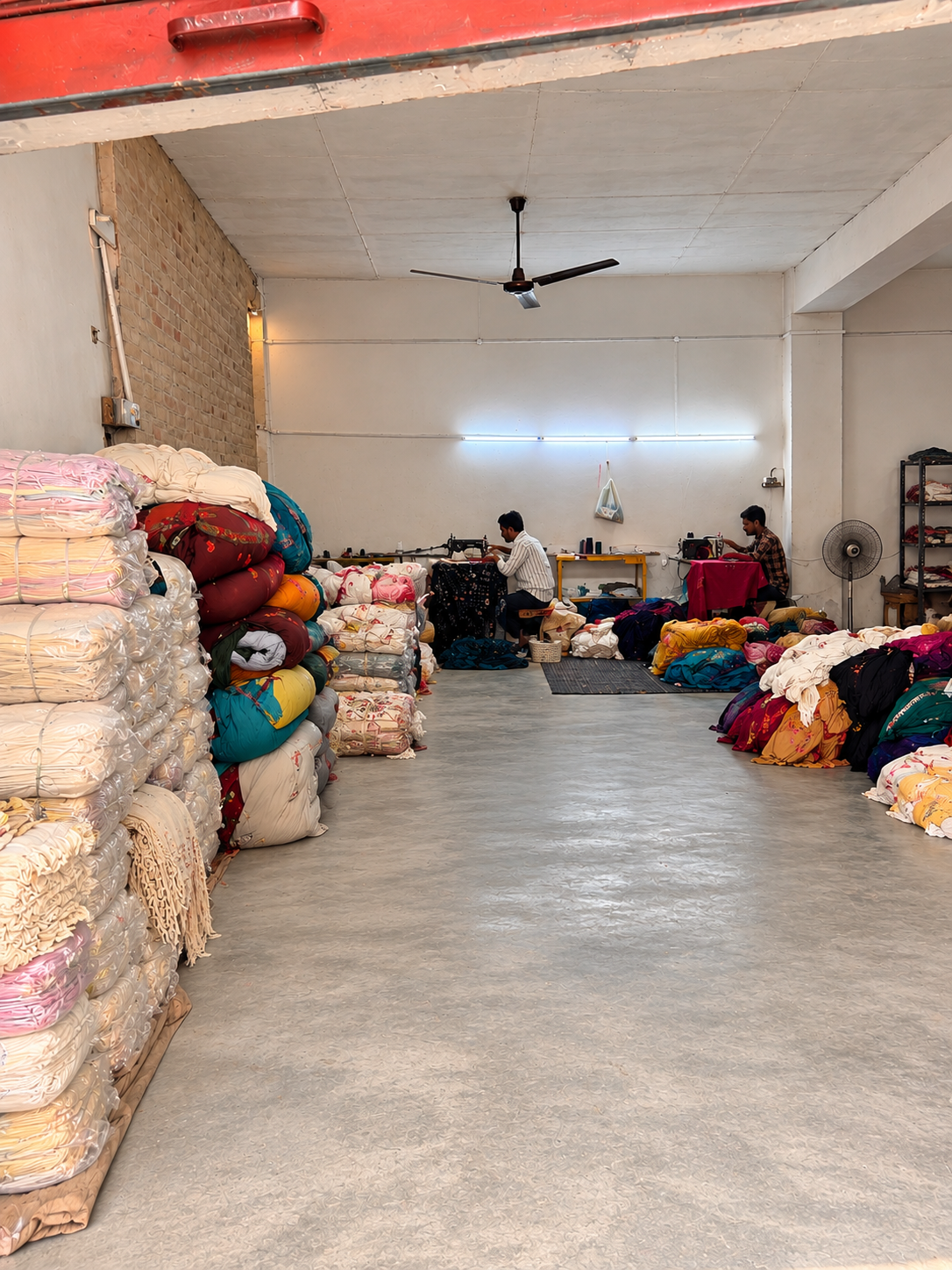 Shawl mending workshop with two people at sewing machines and stacks of embroidered fabrics